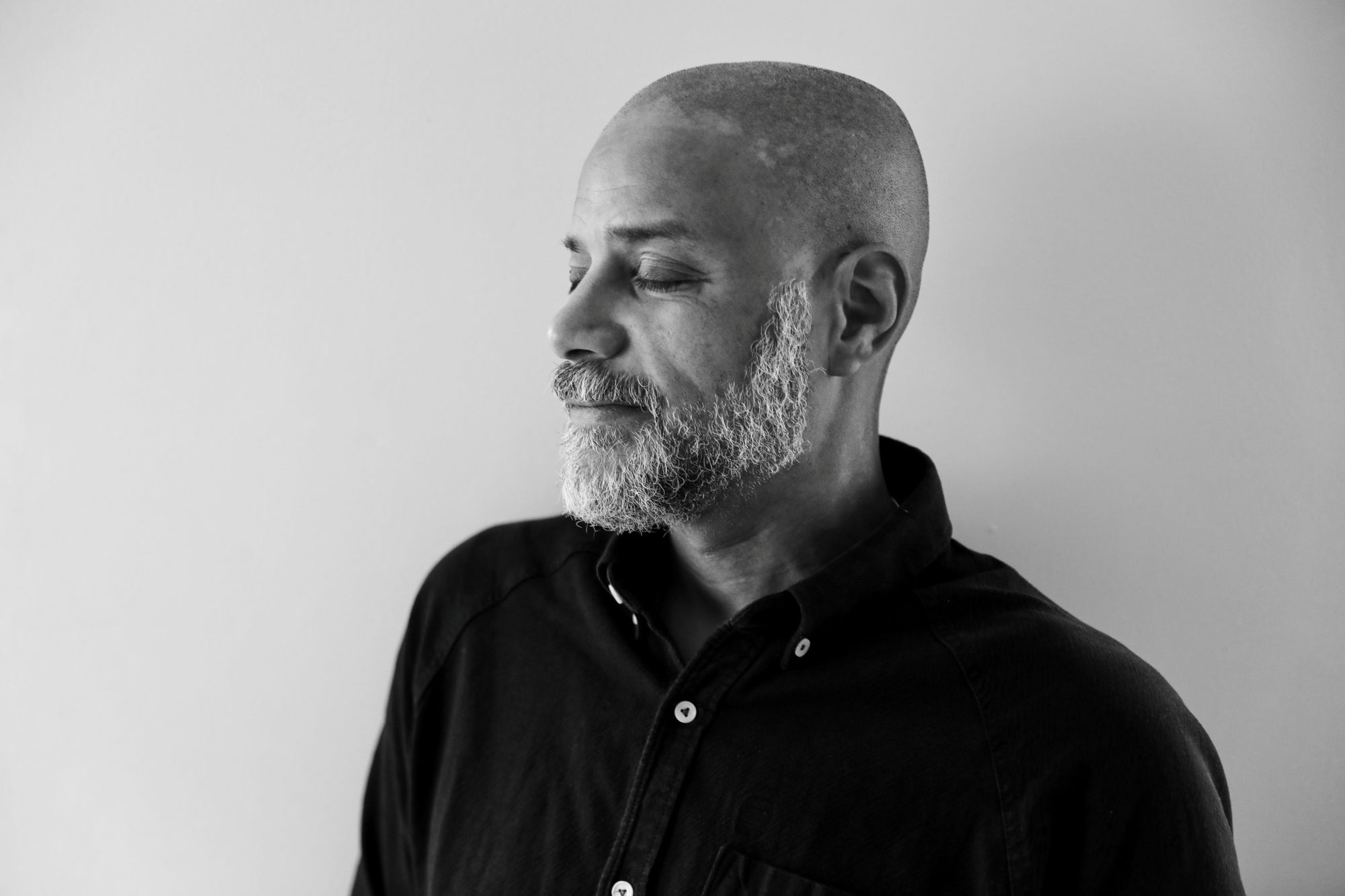 Man with a shaved head and beard wearing a black shirt against a plain background