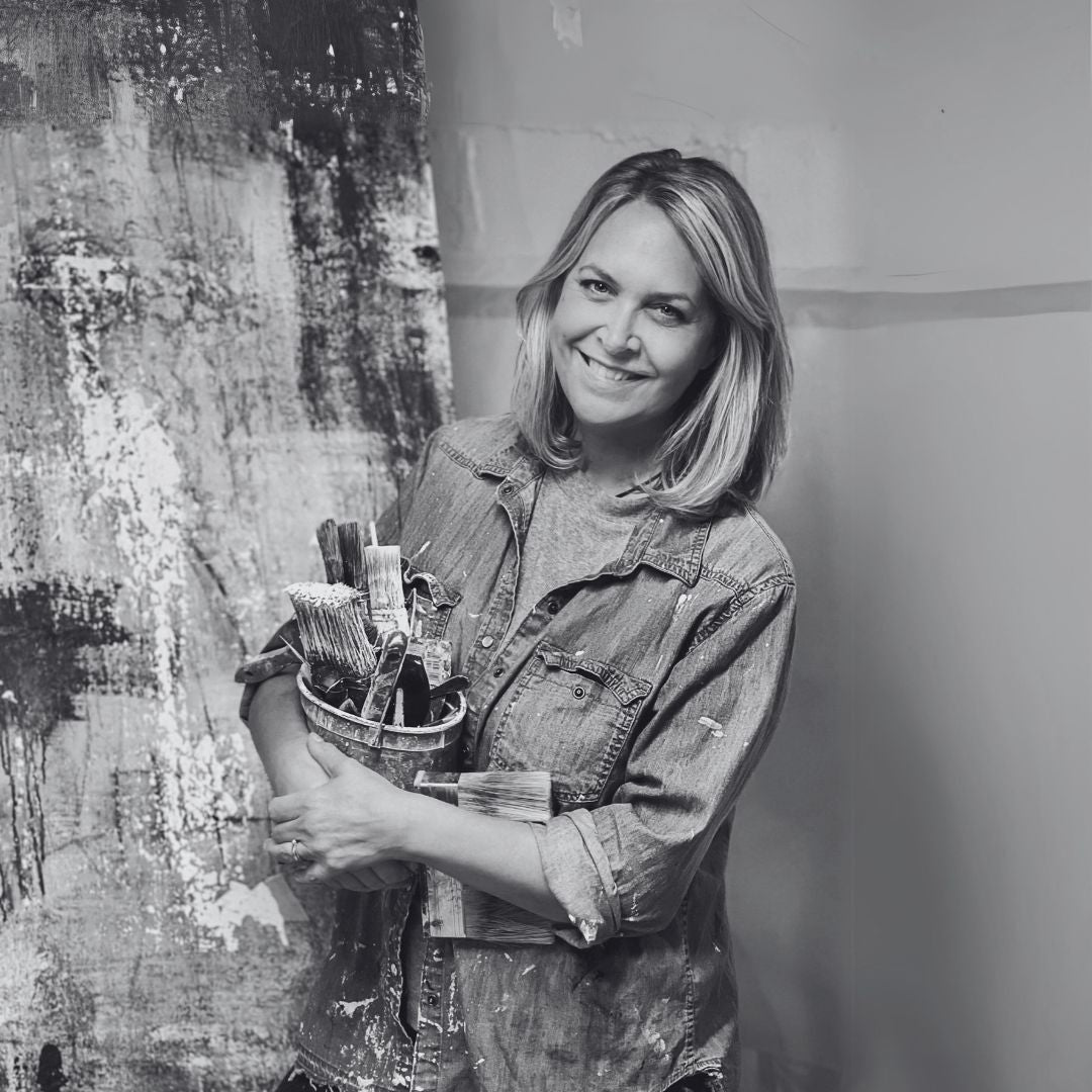 Black and white photo of a woman holding paintbrushes in front of a large artwork