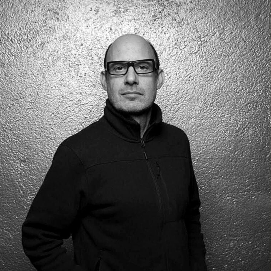 Black and white portrait of a person wearing glasses and a dark jacket against a textured wall.