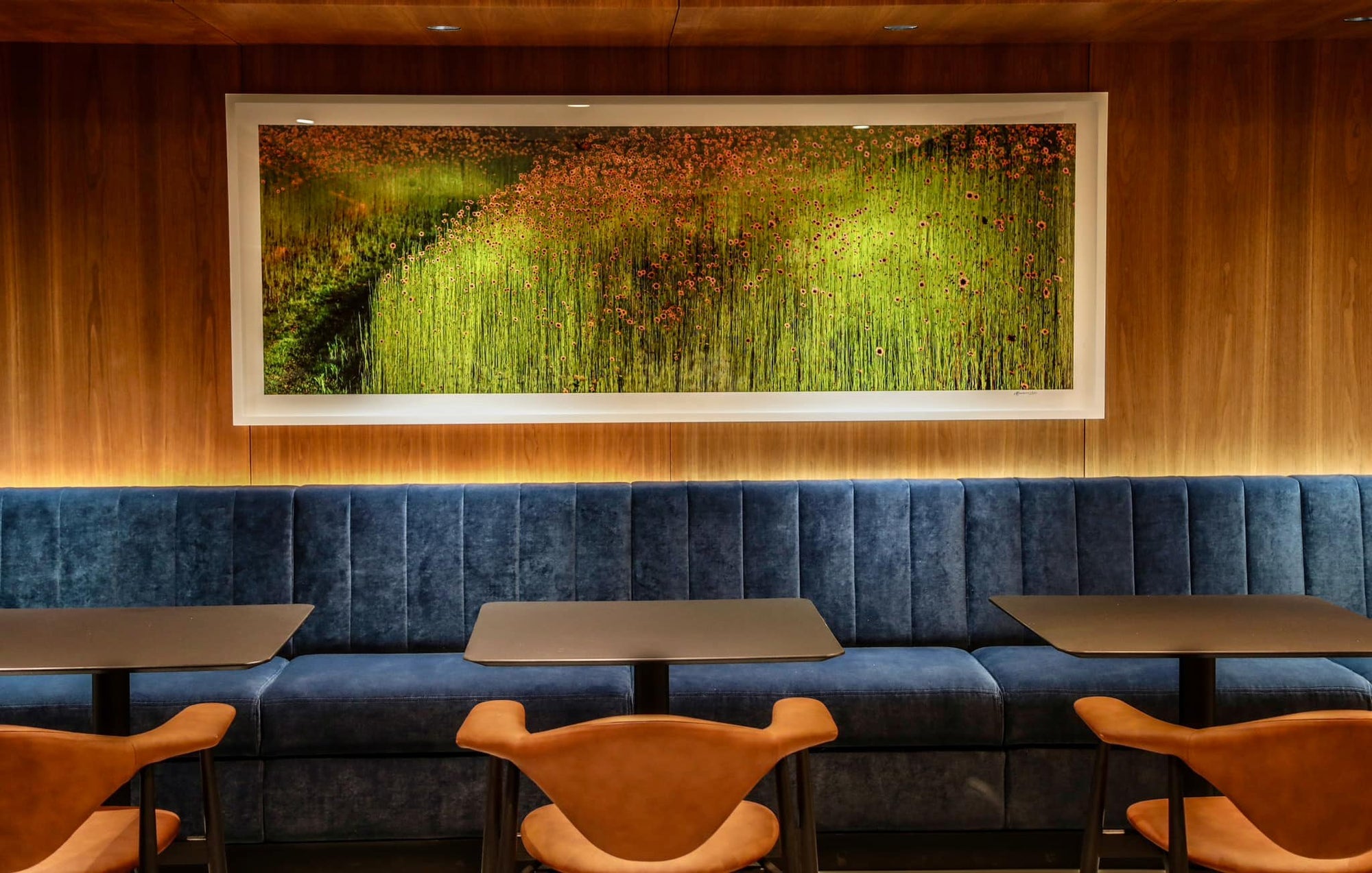 Blue cushioned booth with wooden chairs against a wall with a large framed print.