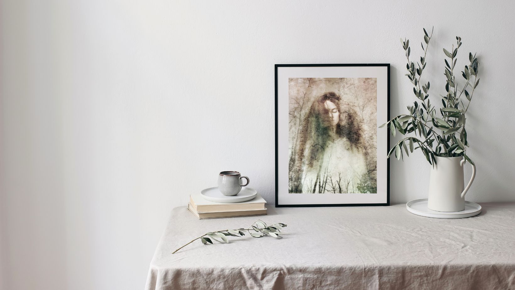 Framed artwork on a table with a cup, books, and a plant.
