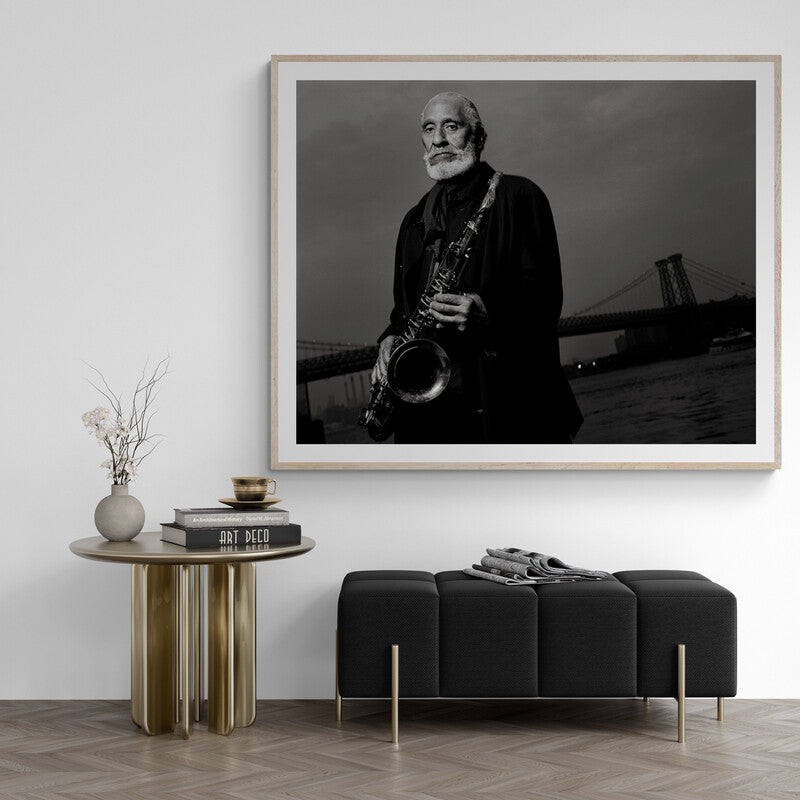 Framed black and white artwork of a man playing a saxophone in a room with a table and bench.