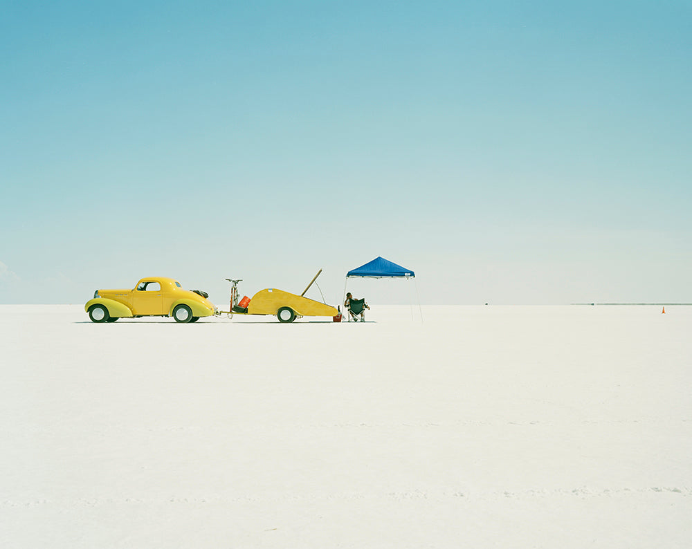 Two yellow cars on a salt lake with a person and a blue tent in the background.