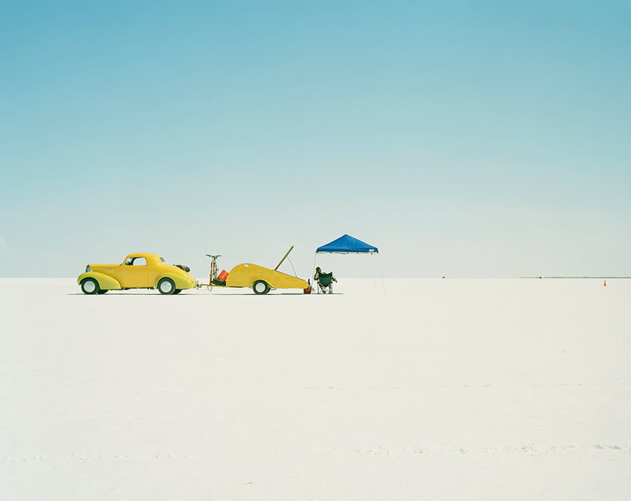 Two yellow cars on a salt lake with a person and a blue tent in the background.