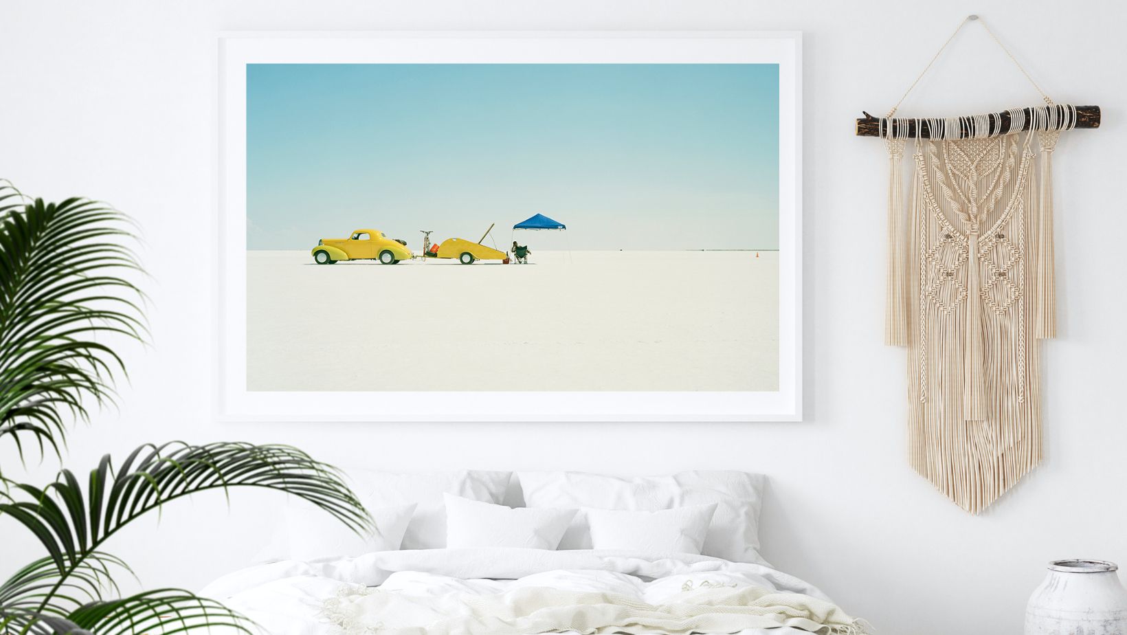 Framed artwork of two yellow cars and a blue umbrella on a white background, hanging above a bed in a room with a macrame wall hanging.