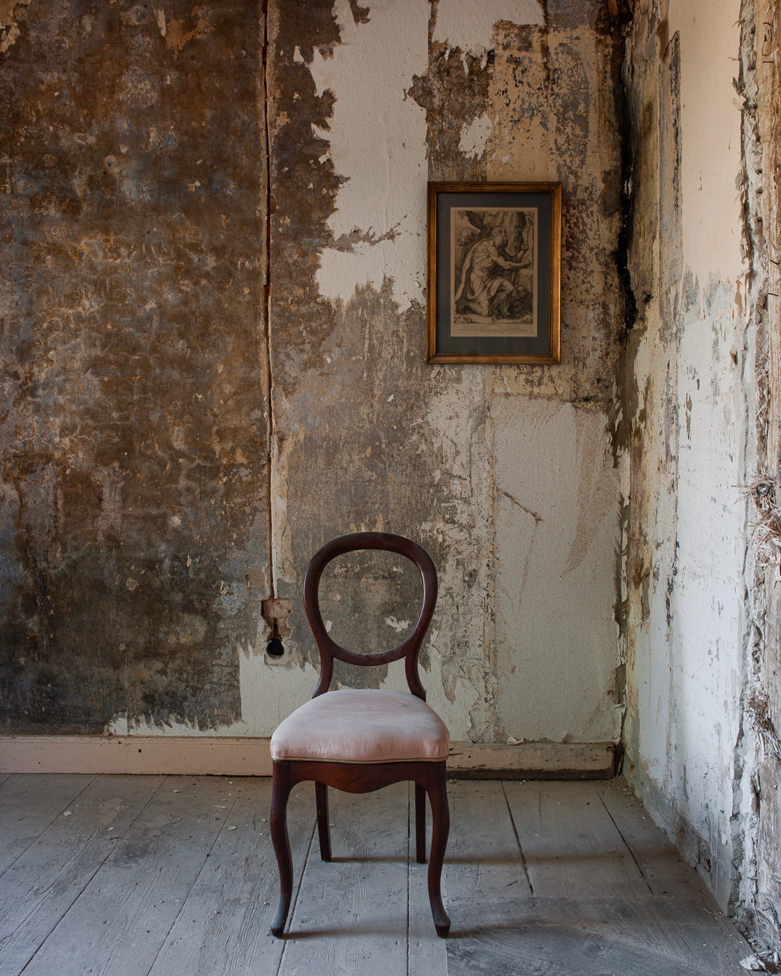 An old chair stands in a room with peeling walls and a framed picture.
