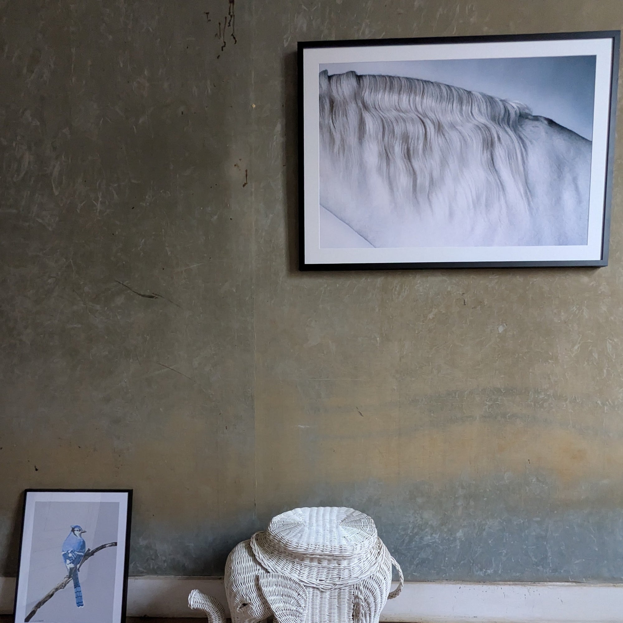 Framed artwork on a textured wall with a wicker stool below.