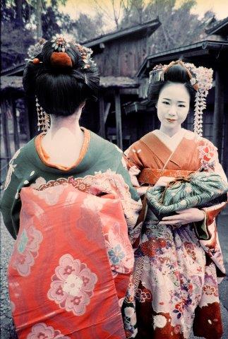 Two women in traditional Japanese kimonos standing outdoors with wooden buildings in the background.