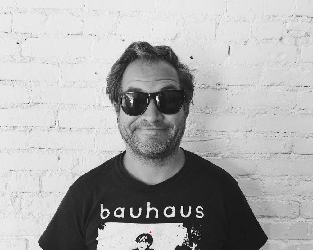 Person wearing a black t-shirt with 'bauhaus' design against a white brick wall.