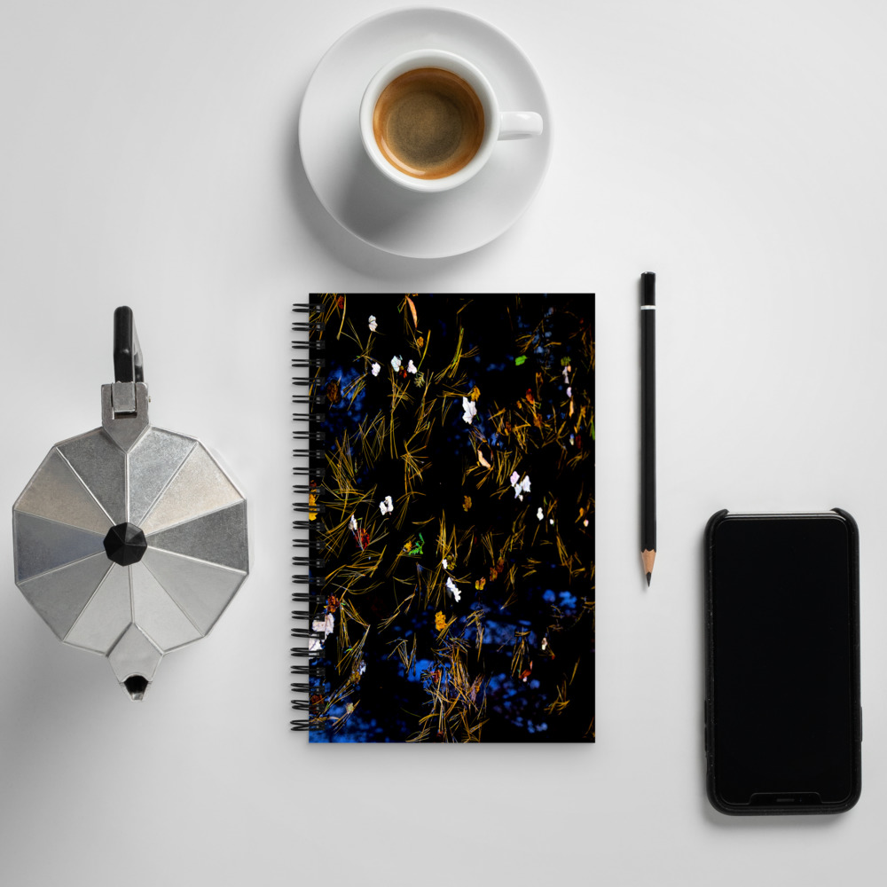 Spiral notebook with a photo of a pond covered with flower petals and leaves on its cover next to a coffee cup and cell phone