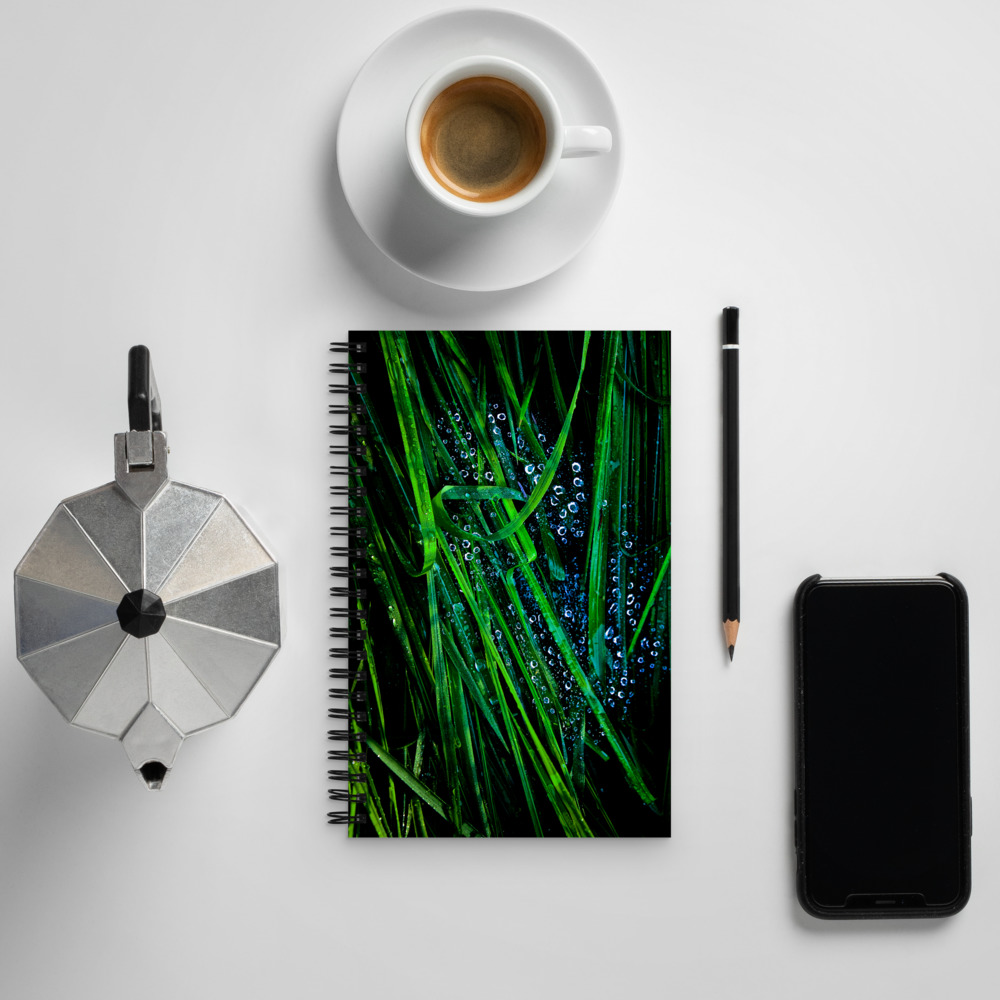 Spiral notebook with a photo of blades of grass covered with water droplets next to a cup of coffee and cell phone
