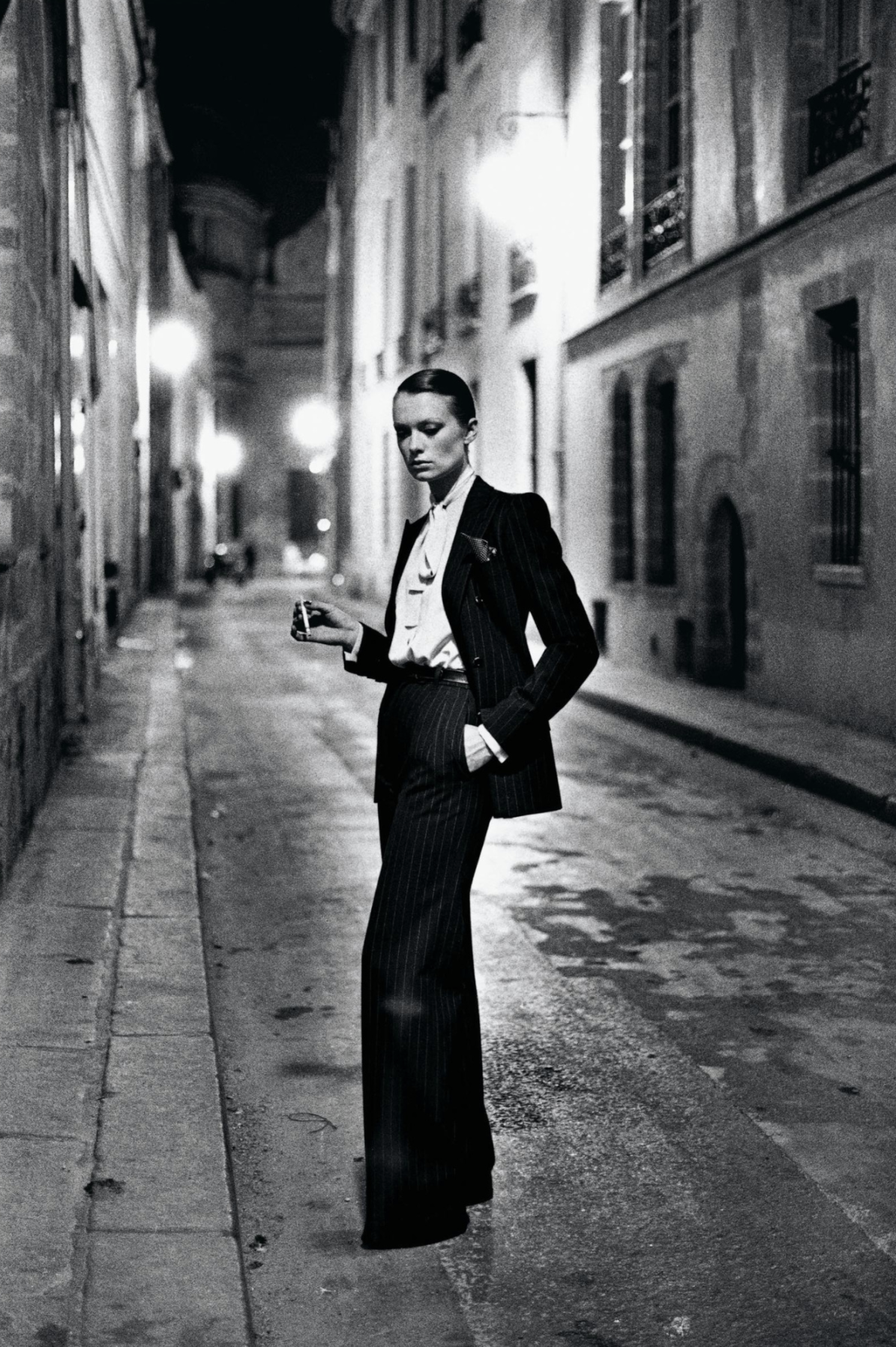 Woman in a tuxedo standing in a street at night, smoking a cigarette