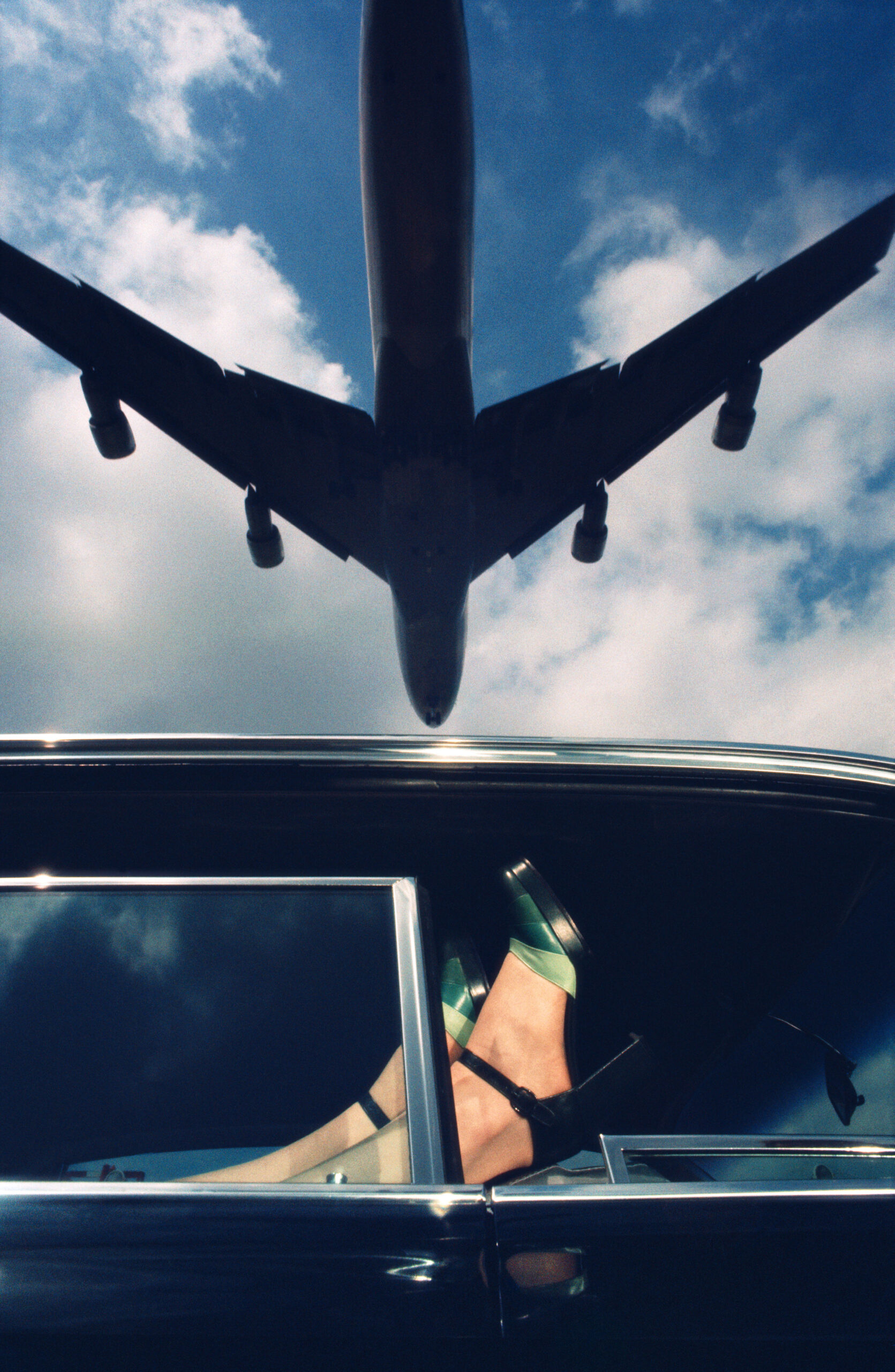 Outside of a car, we see a woman's feet up in the window while a plane flies overhead