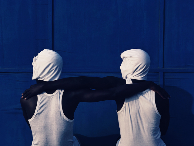 Two black men in white tshirts, their head covered with white fabric, standing in front of a blue wall