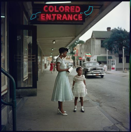 A Black woman with her child standing on a sidewalk near a "Colored Entrance" neon sign