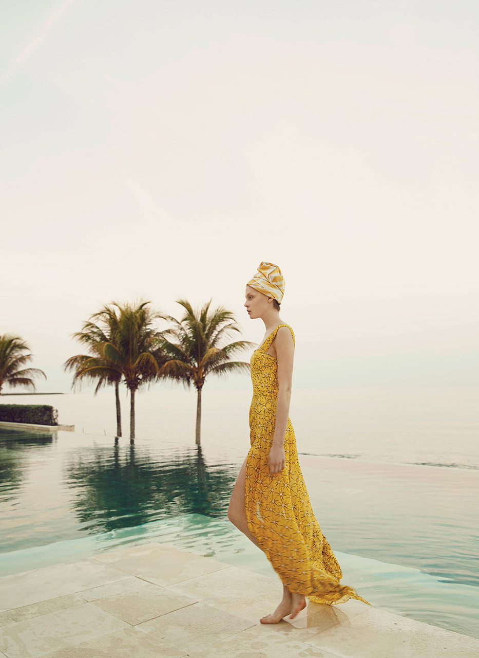 Woman in a long dress, walking by a pool, with palm trees in the background