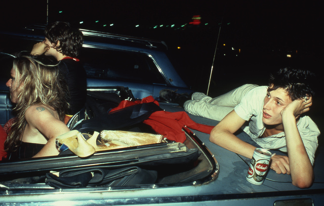 A young man laying on the trunck of a convertible car watching a movie at a drive-in, while his friends sit in the car