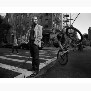 Miguel Zennon holding his saxophone, standing at a street corner in New York City, with a kid doing stunts on his bike in the background