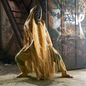 A mysterious figure shrouded in a traditional African costume and hidden by a mask stands in an abandoned building