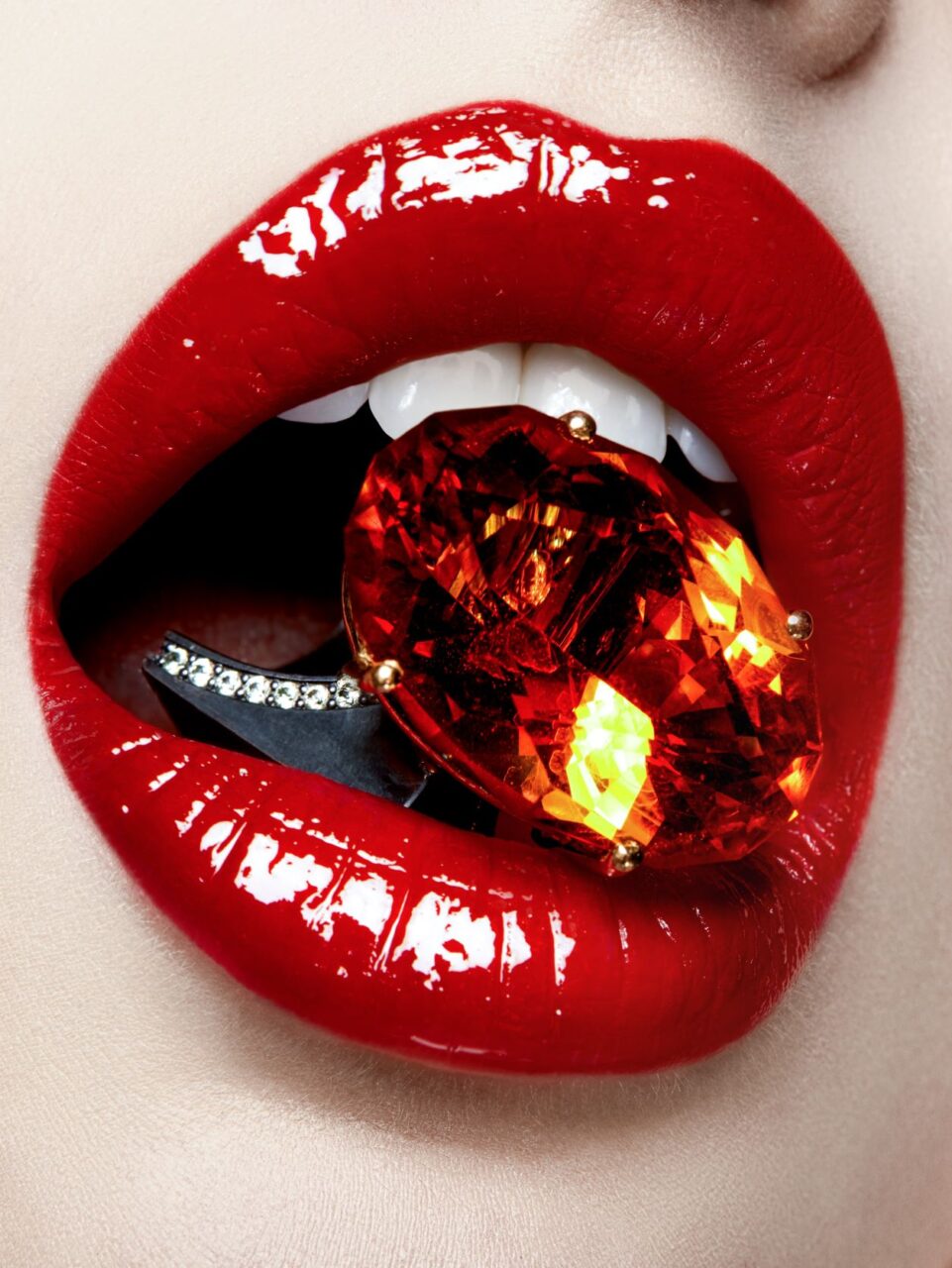 Close-up of a woman's mouth with red lipstick, holding a precious gem between her teeth