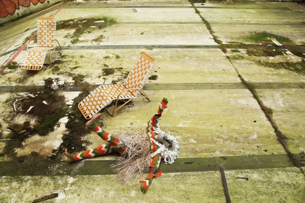 A mysterious figure in an African costume and mask lies down on the floor of an abandoned building