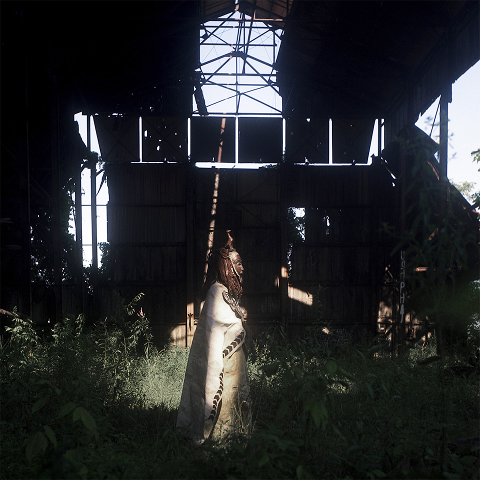 A mysterious figure in an African costume and mask stands in the middle of an overgrown abandoned building