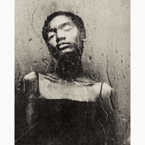 Young Black woman with her eyes closed, standing by a wall, as seen through a rainy window pane