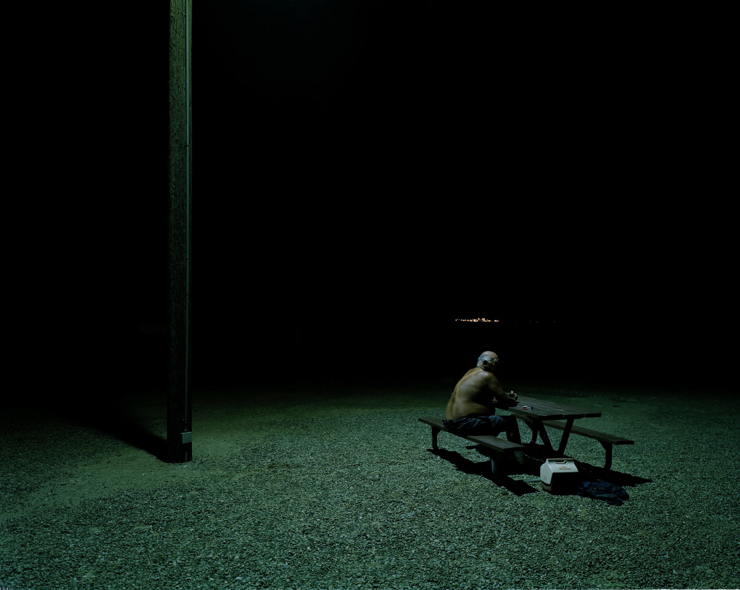 Lone person, seen from the back, sitting at a picnic table in a park at night