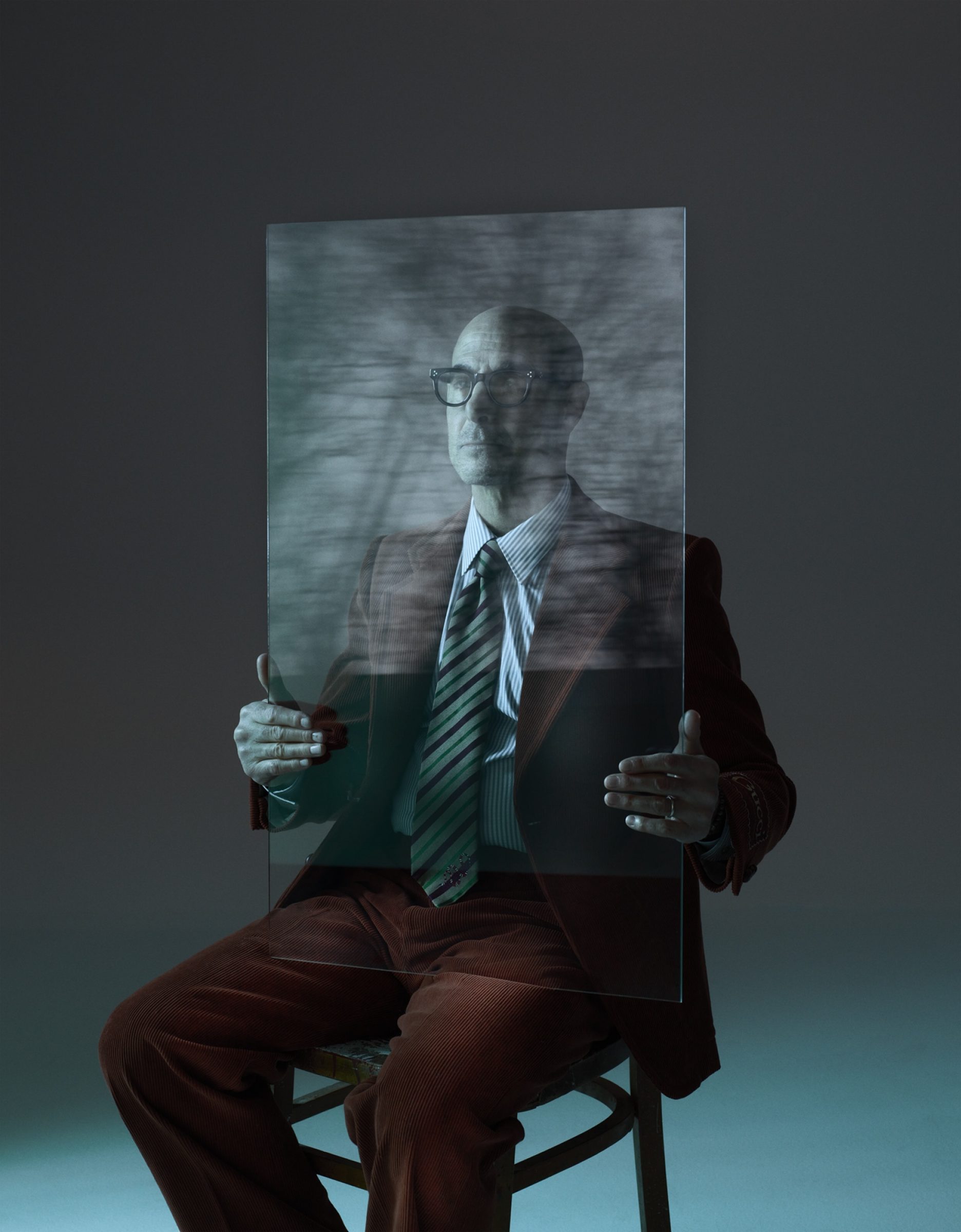 Actor Stanley Tucci sitting on a chair in a dark room, holding a glass panel in front of him