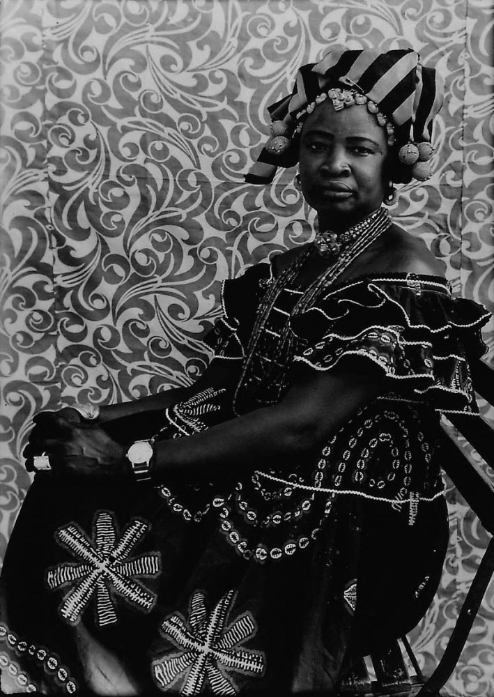 Formal portrait of a Malian woman, dressed in traditionnal dress and headdress, sitting regaly in front of a richly patterned African fabric