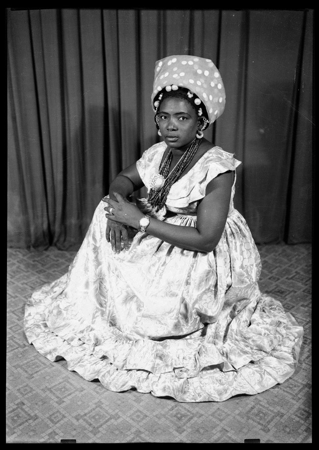 Portrait of a Malian woman, with her traditional dress laid down around her, sitting down, with a patterned fabric in the background