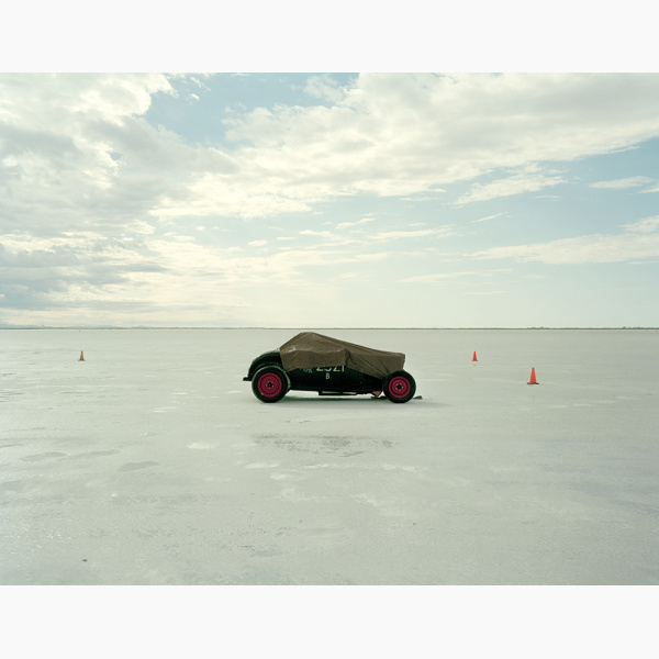 An old racing car stands alone on a salt flat with a few traffic cones nearby