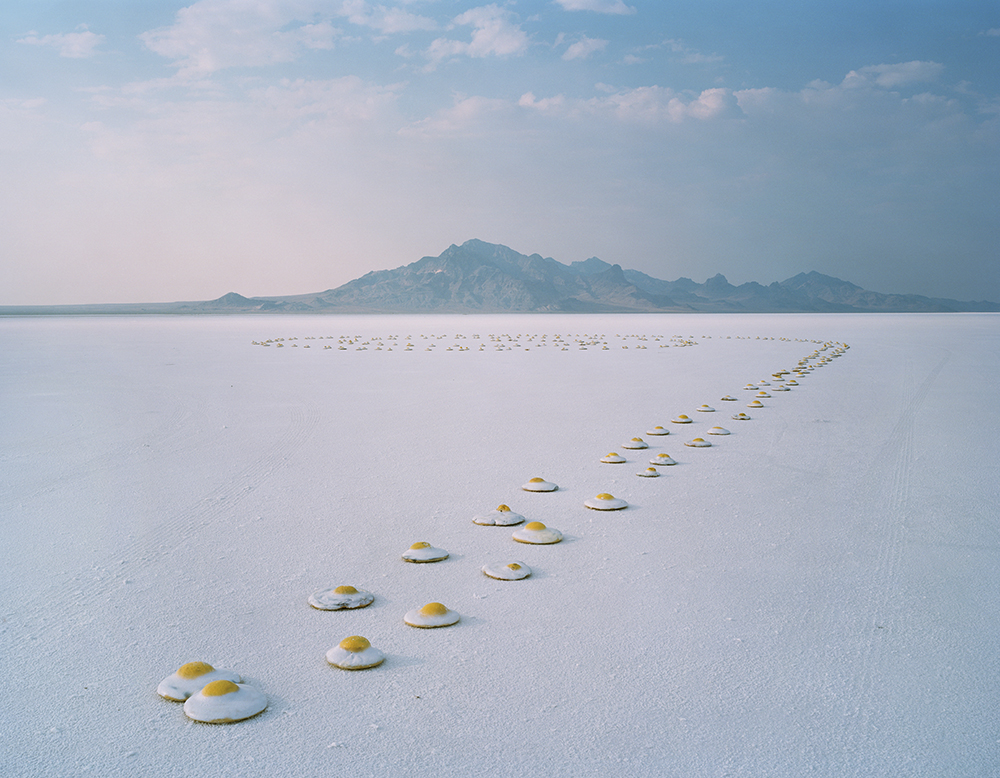 A row of gag plastic poached eggs lays on the ground of a salt flat with mountains in the distance