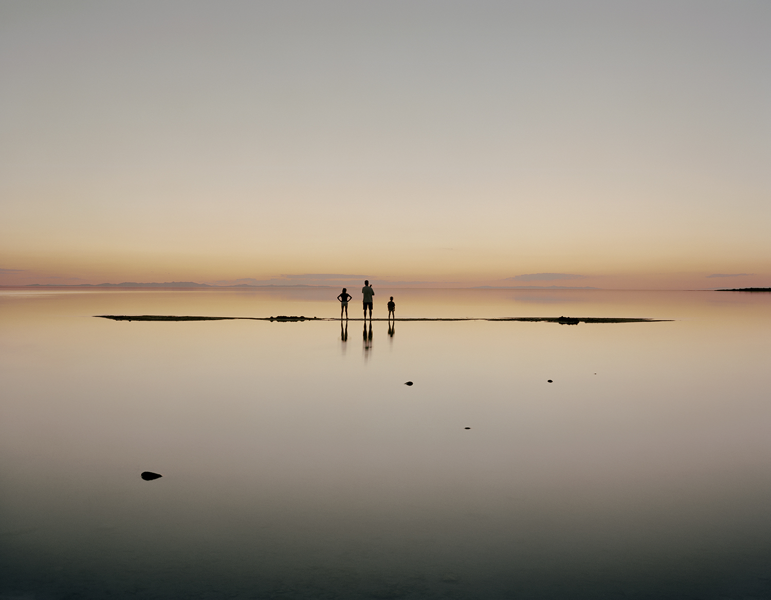 A man with his two kids stands on a raised salt bed in the middle of a lake at sunset