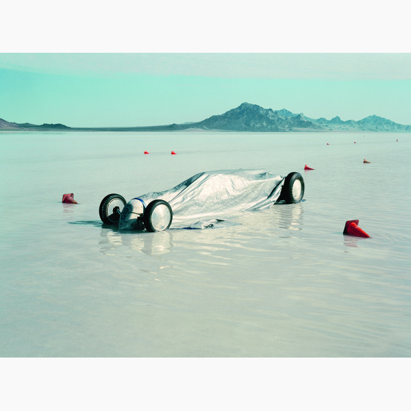 A racing car is covered by a silver cloak in the middle of a large pool of water on a salt flat