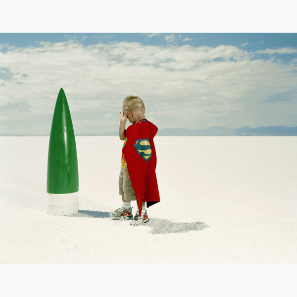 A little boy with a Superman cape stands in front of the tip of a rocket